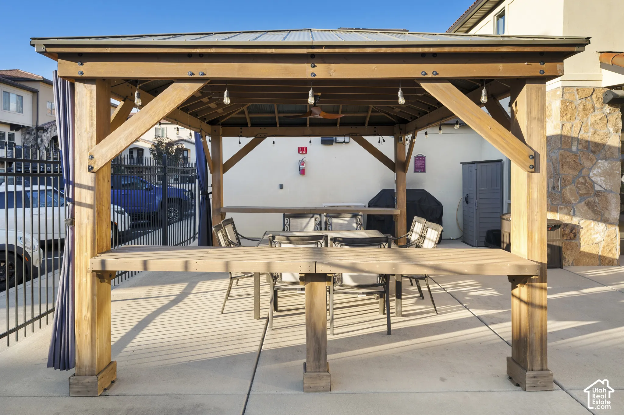 Freestanding wood-frame gazebo with a metal roof, integrated bar-style counter, and ceiling fan, situated on a concrete patio with gas grill