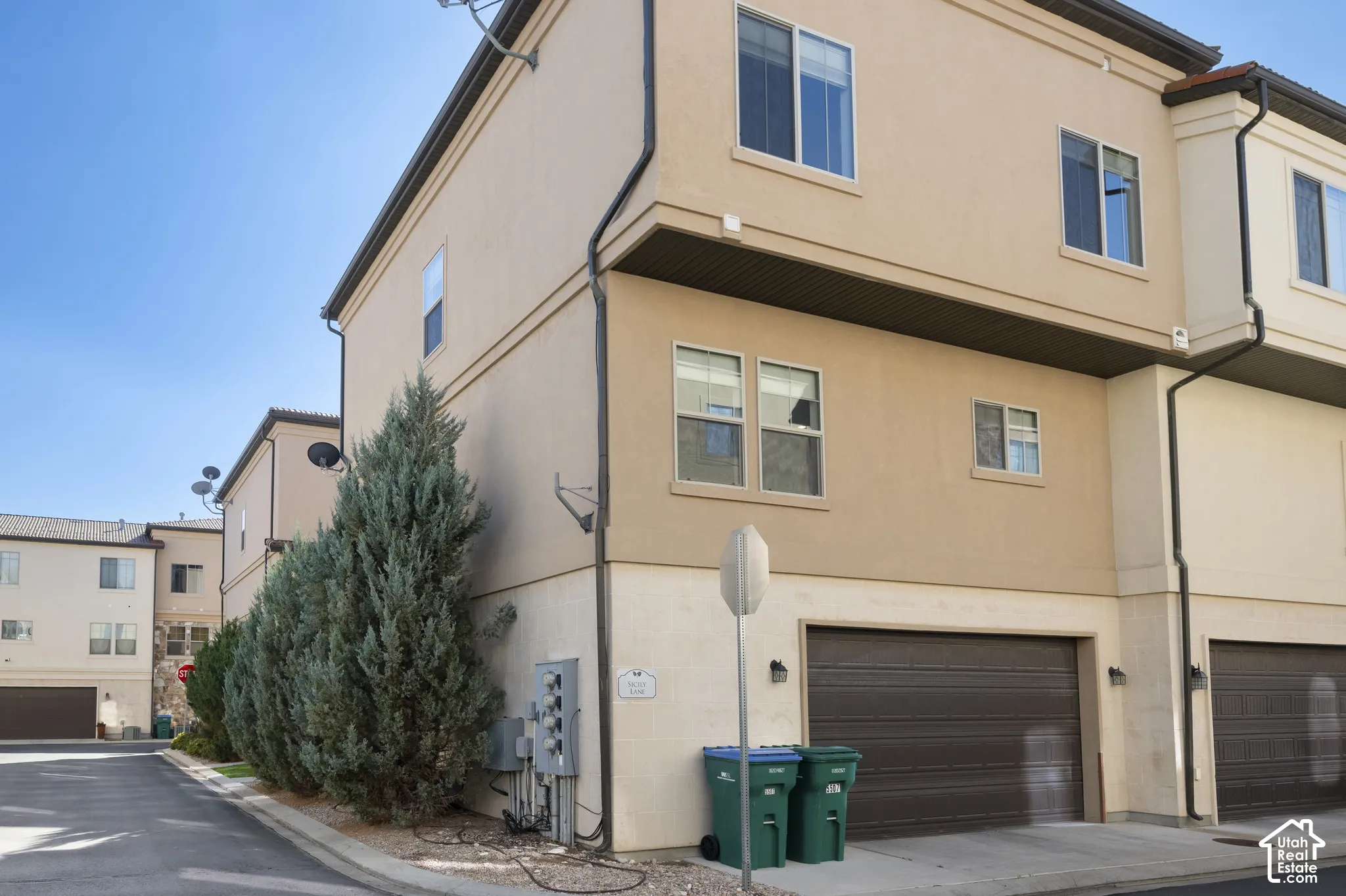 End unit multi-story exterior featuring tan stucco siding, dark-toned sectional 2 car garage door, multiple rectangular windows, and a built-in exterior light fixture