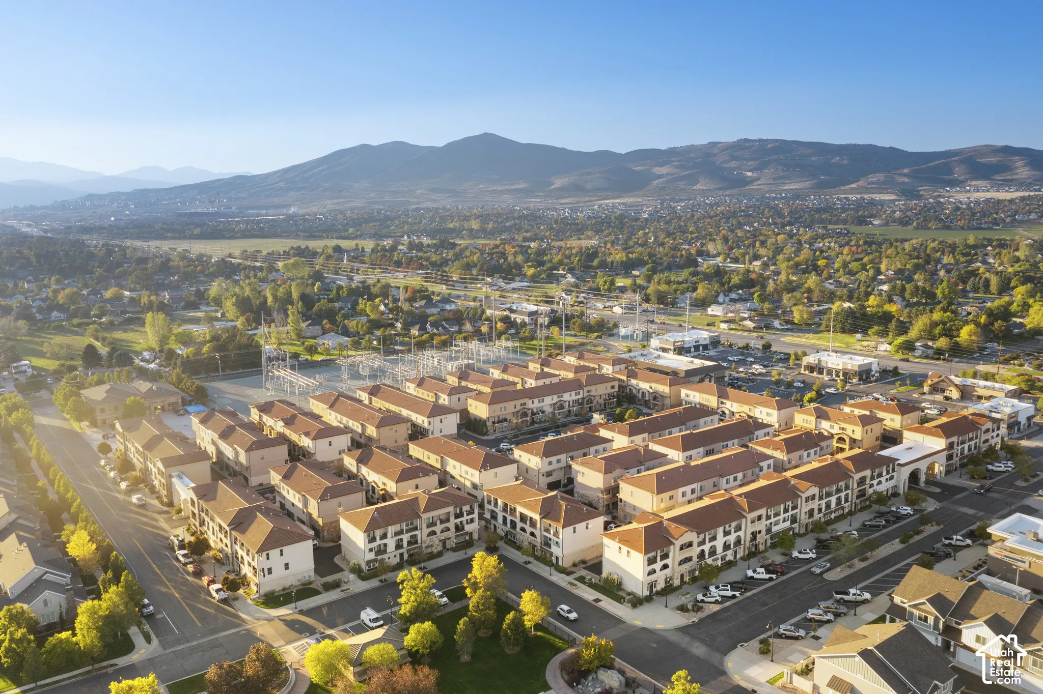 Aerial perspective of a multi-unit residential complex featuring Mediterranean-style architecture, red tile roofing, light-toned stucco exteriors, paved street access, and a mountain range backdrop
