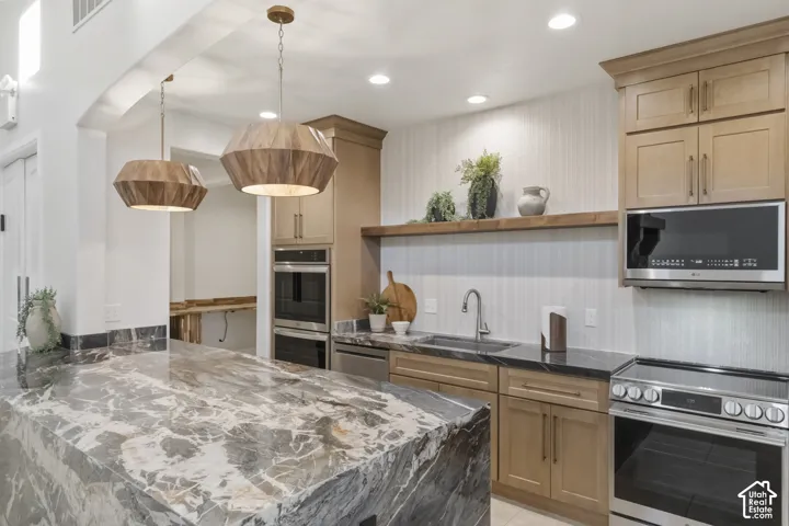Kitchen featuring an expansive stone-topped island, light-toned custom cabinetry, built-in stainless steel three ovens, a stainless steel range, and geometric wood-finish pendant lighting and a butler kitchen for preparation in "back of house"