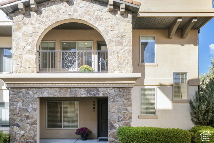 Stucco exterior featuring stone masonry accents, an arched entryway, a second-story balcony with decorative iron railing, and corbel detailing