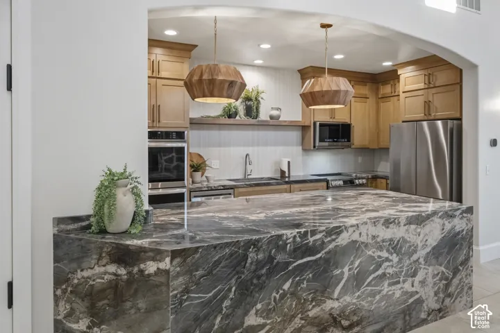 Clubhouse kitchen featuring a large waterfall-edge stone island, natural wood cabinetry, stainless steel appliances, recessed lighting, and geometric wood-finish pendant fixtures
