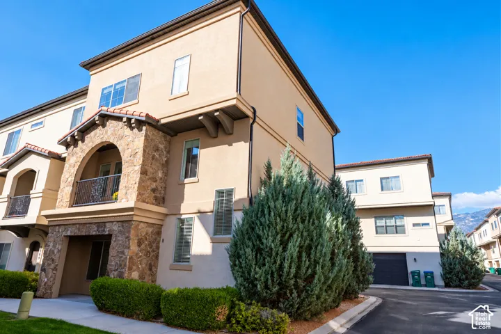 Multi-level stucco exterior featuring stone masonry accents, an arched balcony with metal railing, decorative corbel details, and a paved driveway