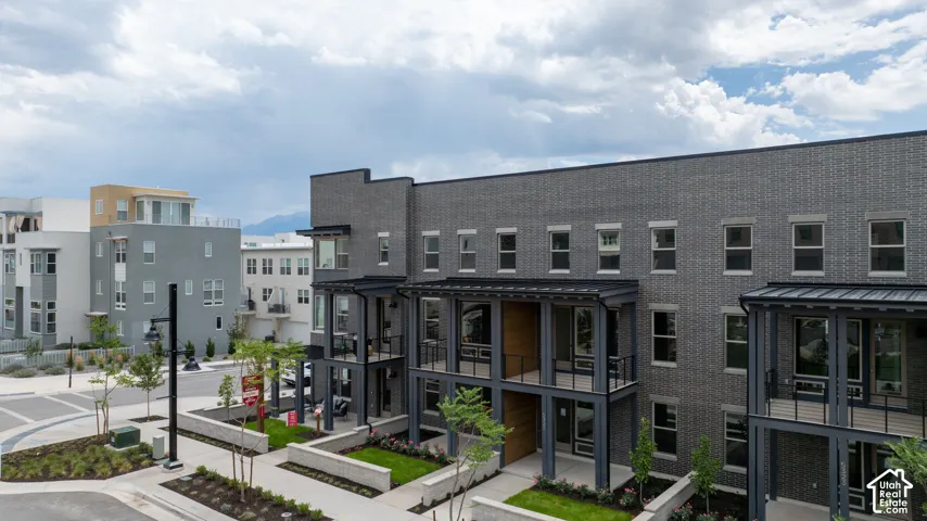 Multi-story exterior featuring dark brick facade, metal-roofed covered balconies, metal structural support columns, and ground-level concrete retaining walls with integrated landscaping
