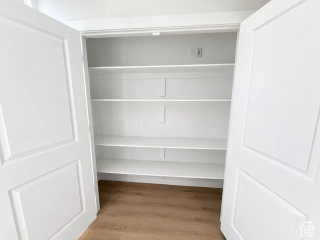 Storage closet featuring double doors, four tiers of white shelving, wood-finish flooring, and electrical outlets