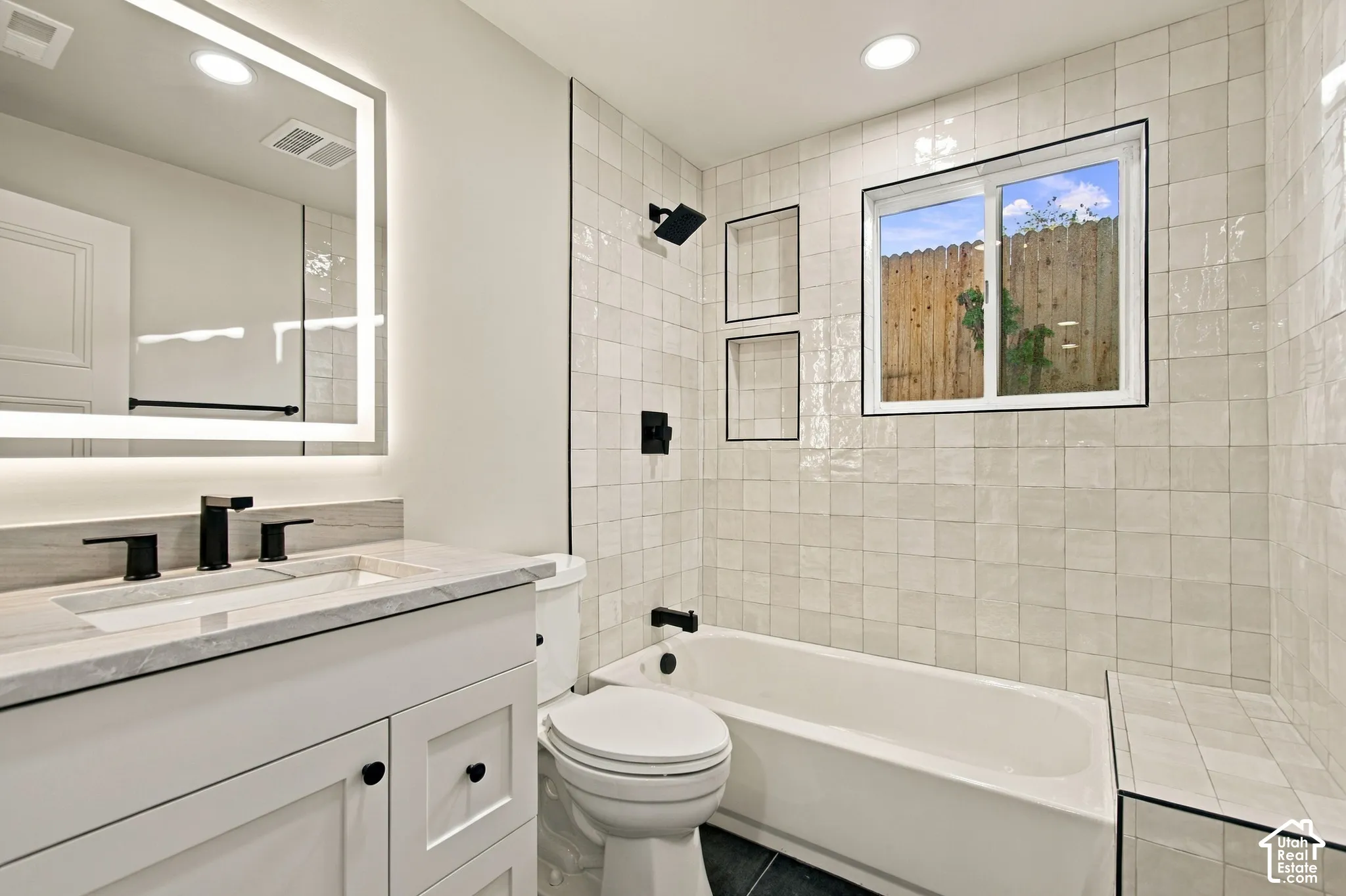 White vanity cabinet with stone-finish countertop, integrated under-mount sink, matte black faucet, wall-mounted LED-backlit mirror, and tile shower surround with dual built-in recessed niches
