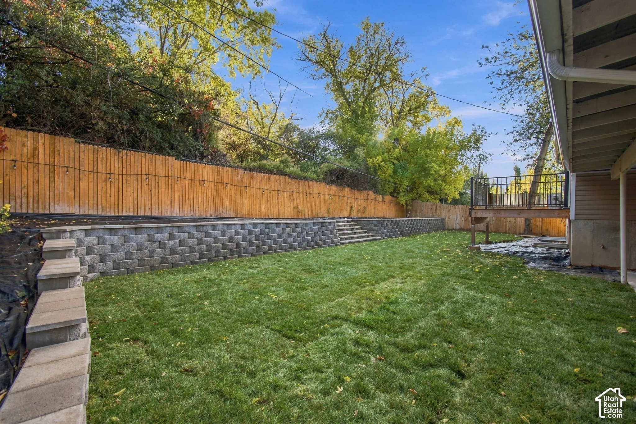 Backyard featuring a tiered stone retaining wall, natural grass lawn, wood privacy fencing, and an elevated deck with metal railing