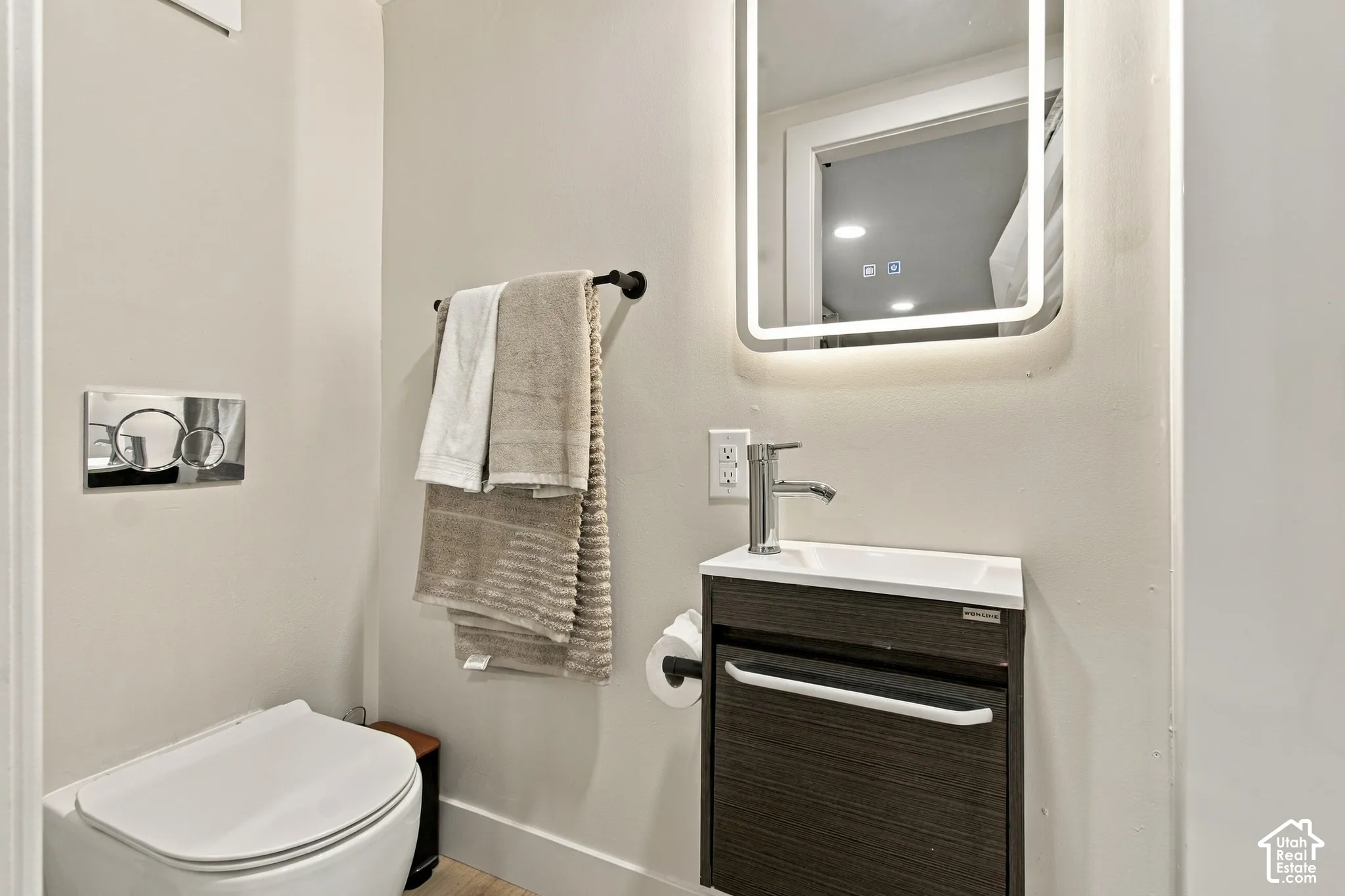 Wall-mounted toilet with a chrome dual-flush actuator, vanity with an integrated sink, polished chrome faucet, and LED-backlit mirror
