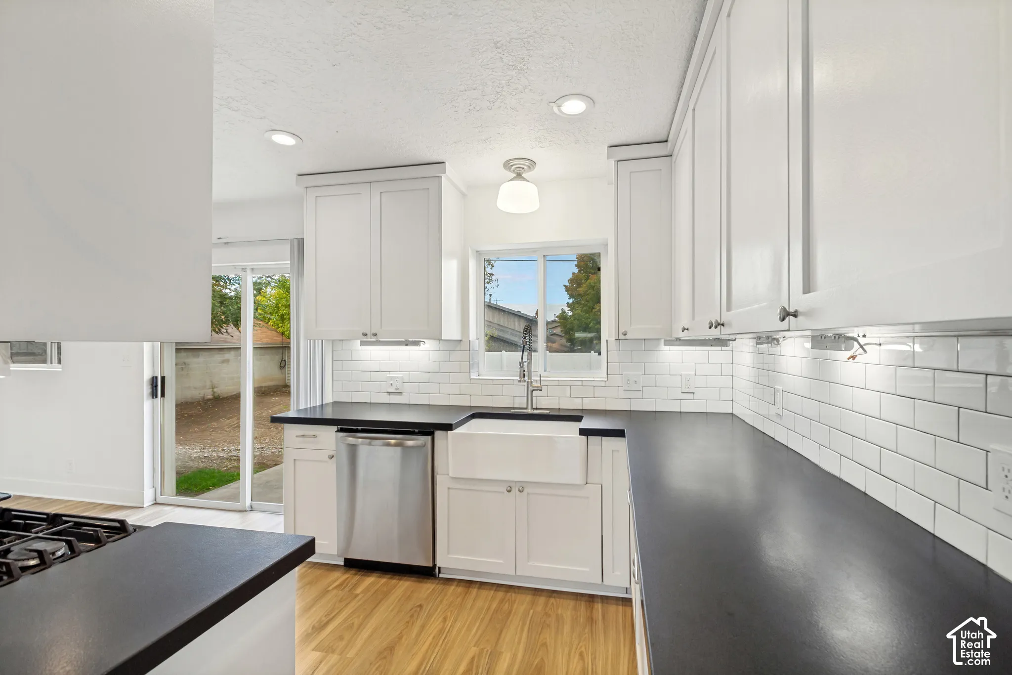 Kitchen featuring white Shaker-style cabinetry, dark countertops, a white farmhouse sink, subway tile backsplash, and wood-finish flooring