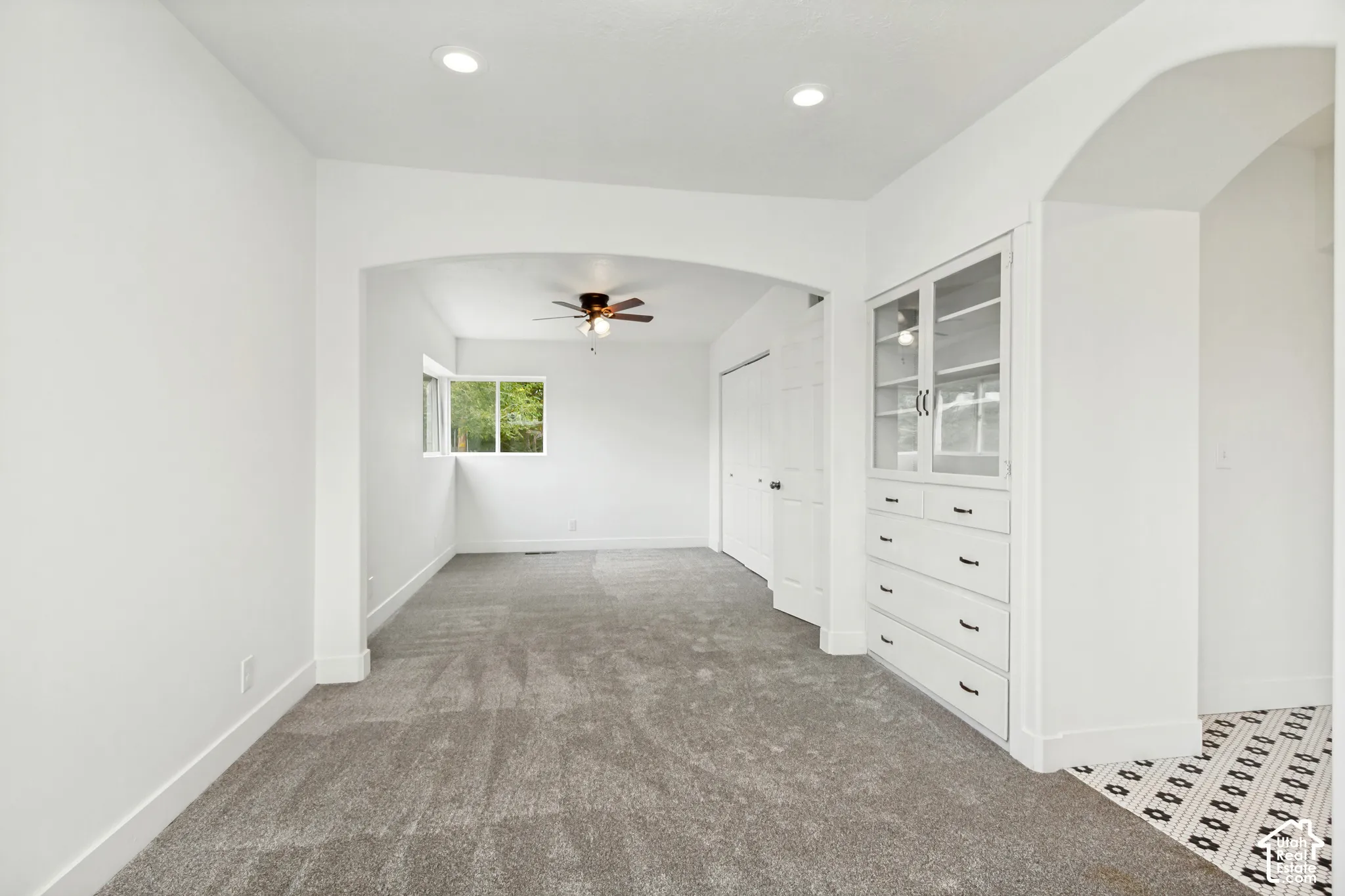 Neutral-toned carpeted flooring, built-in glass-front cabinetry with lower drawers, arched architectural pass-through, recessed ceiling lighting, and ceiling fan fixture