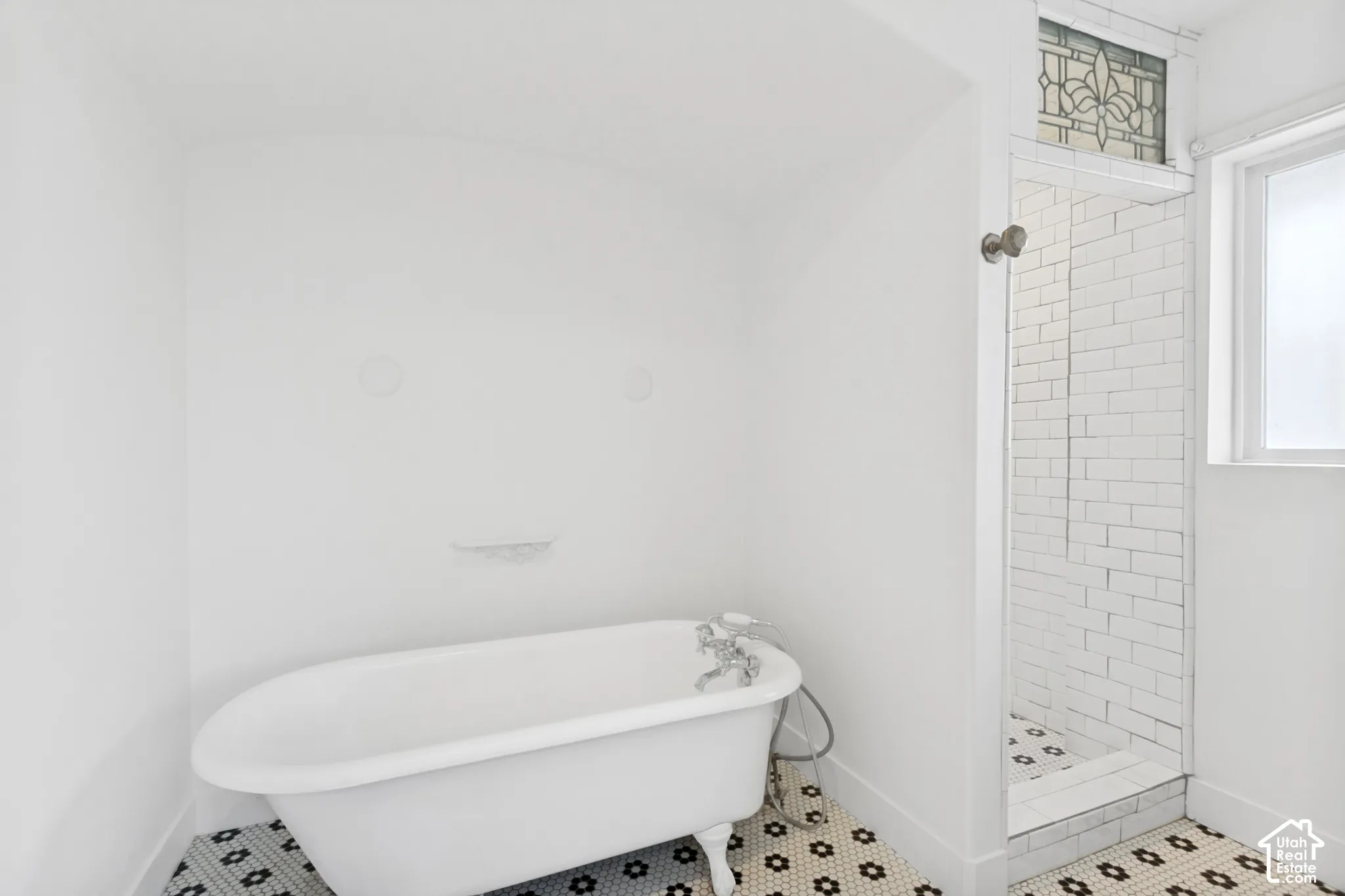 Clawfoot bathtub with metallic hardware, white subway tile shower enclosure, decorative stained glass transom, and patterned mosaic tile flooring