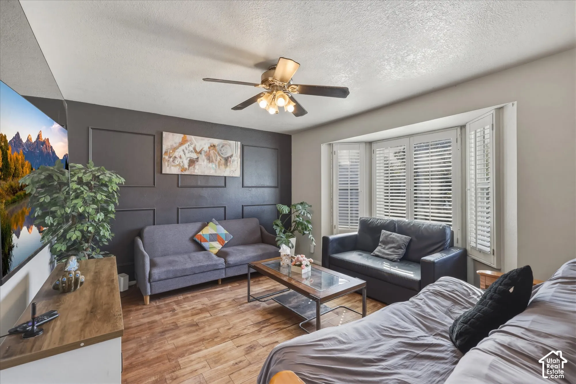 Living area featuring wood-finish flooring, a charcoal-colored accent wall with applied panel molding, a central ceiling fan with multi-light fixture, and a bay window equipped with interior shutters