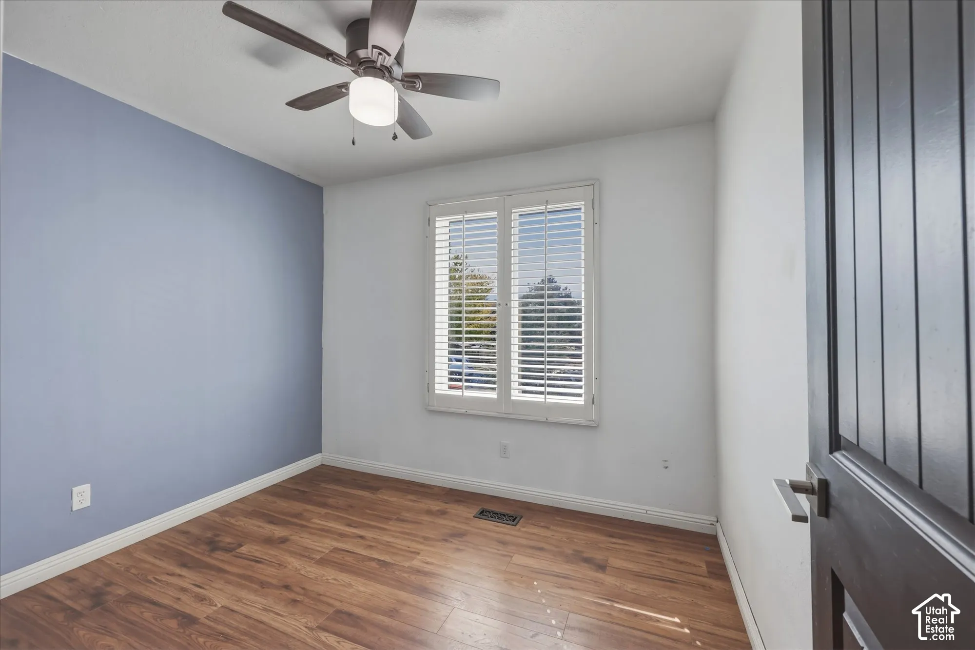 Room featuring wood-finish flooring, accent wall, ceiling fan with integrated lighting, and plantation-style window shutters