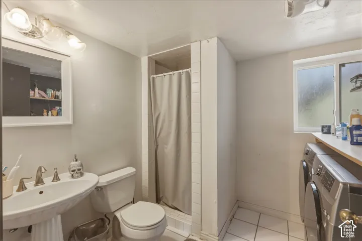 Pedestal sink with metallic faucet, white-tiled shower enclosure, ceramic floor tiling, and dedicated laundry area with built-in shelving