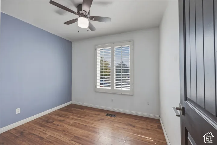 Room featuring wood-finish flooring, accent wall, ceiling fan with integrated lighting, and plantation-style window shutters