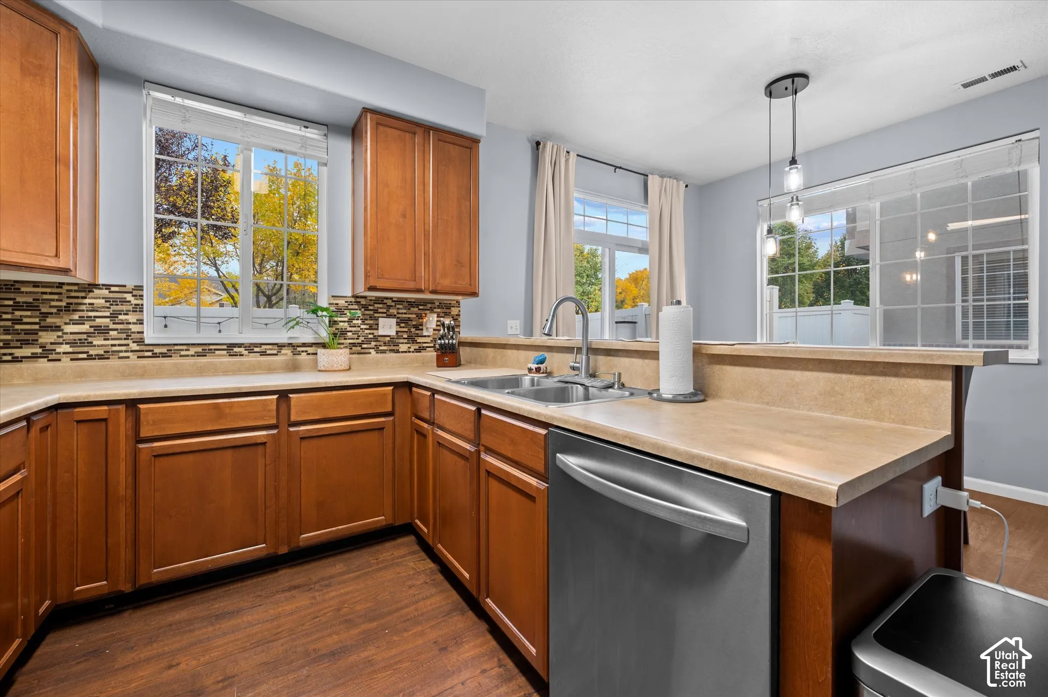 Kitchen featuring wood cabinetry, solid surface countertops, a mosaic tile backsplash, stainless steel dishwasher, and multi-light pendant fixture