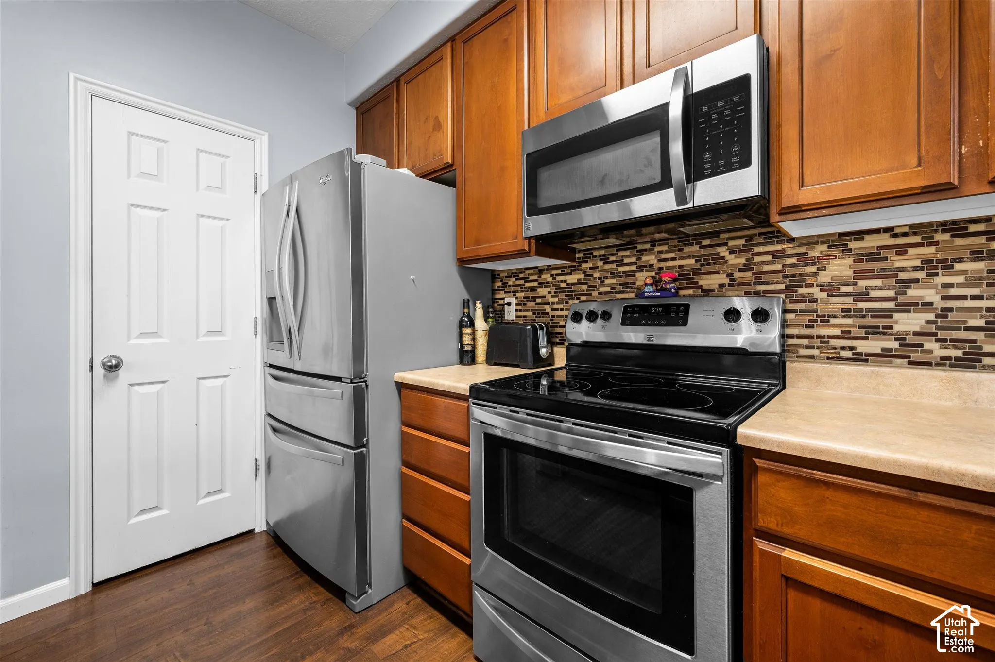Kitchen featuring stained wood cabinetry, stainless steel appliances, multi-tonal mosaic tile backsplash, solid-surface countertops, and wood-finish flooring
