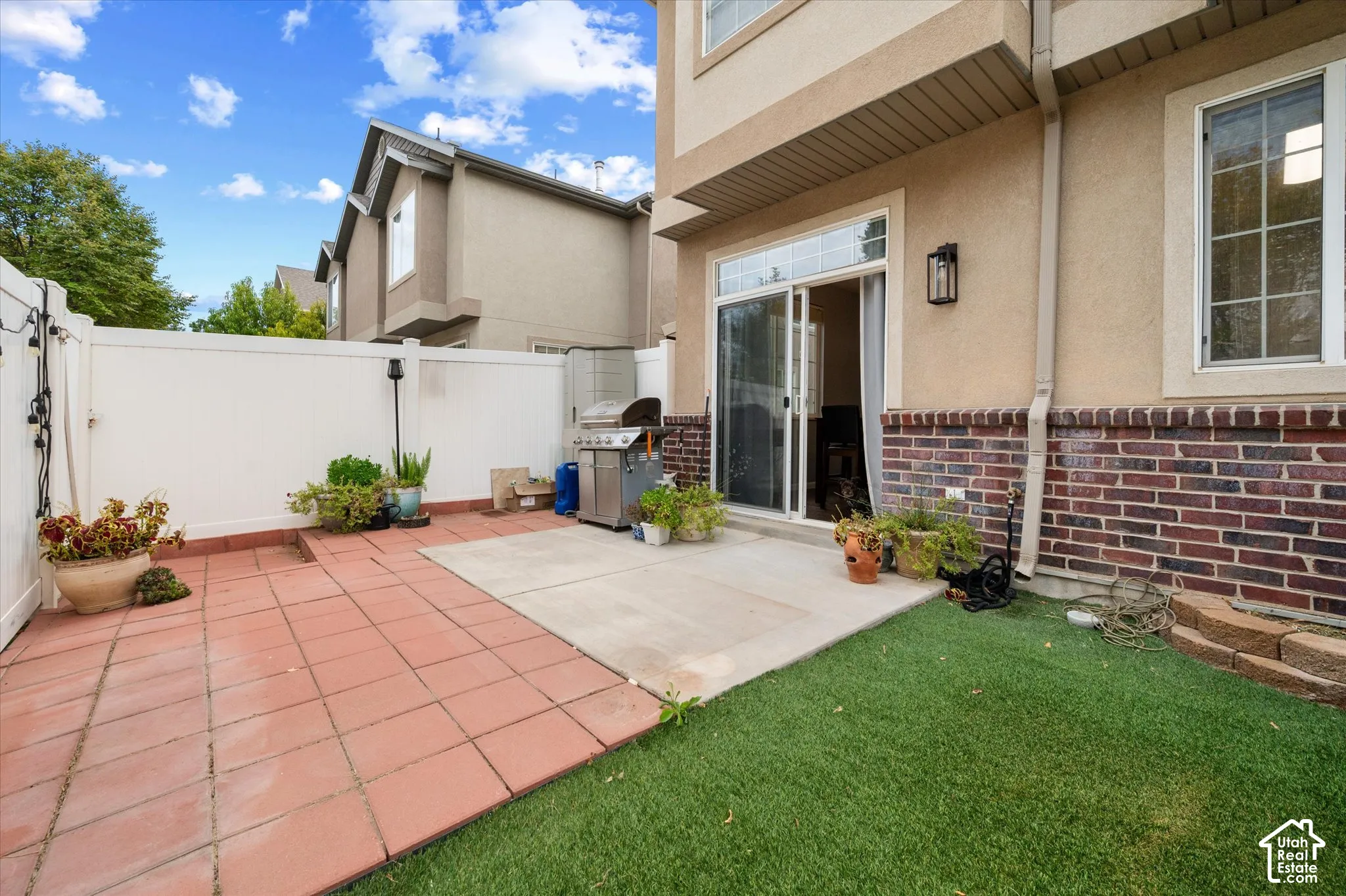 Exterior patio featuring red tile pavers, a concrete slab, artificial turf, and a white privacy fence
