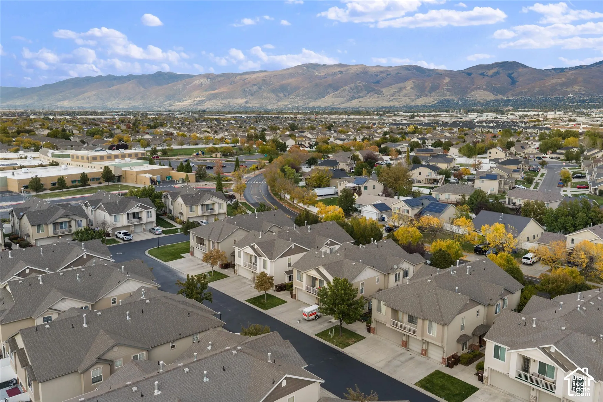 Aerial perspective of a multi-unit residential community featuring asphalt roadways, shingled roofing, mature deciduous trees, and a mountain range backdrop