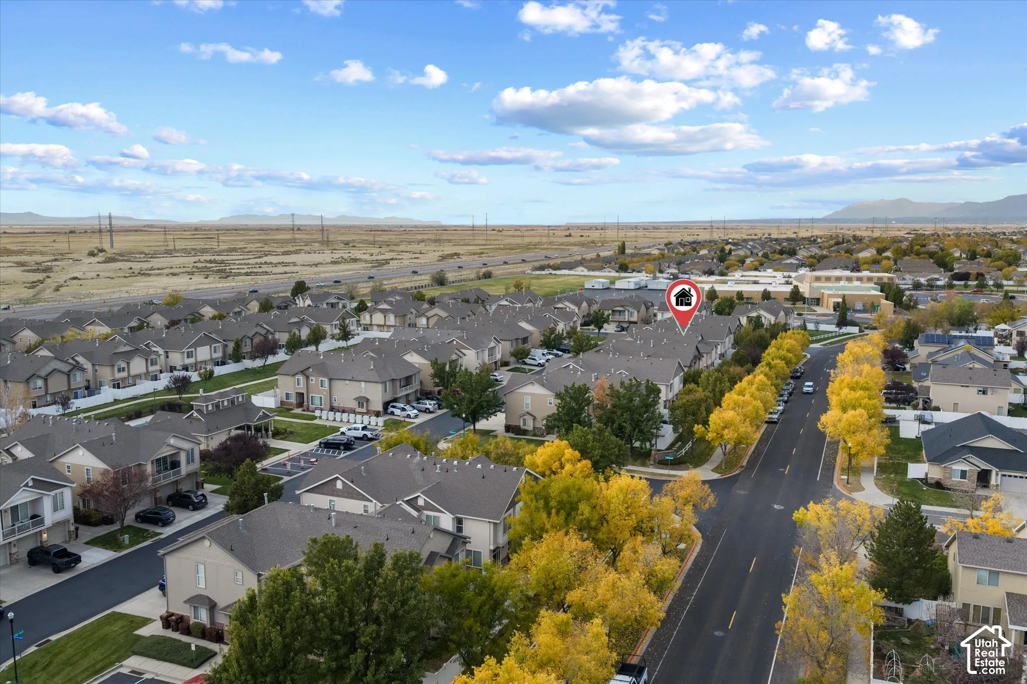 Aerial perspective of a multi-unit residential community featuring asphalt roadways, mature deciduous trees, and surrounding open terrain