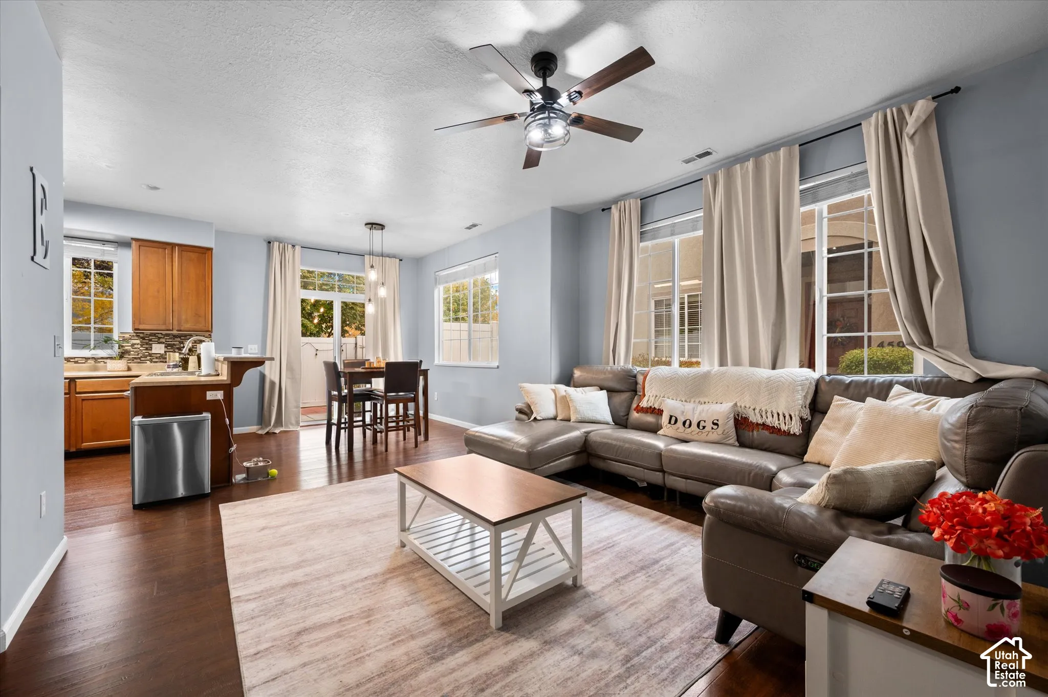 Open floor plan featuring wood-finish flooring, a ceiling fan with integrated lighting, a kitchen breakfast bar, and multiple large windows