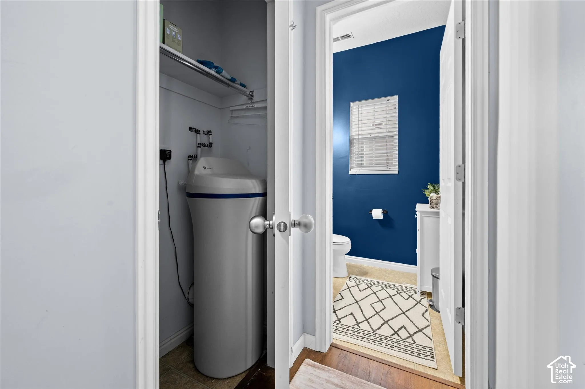 Utility closet featuring a water softener system and integrated shelving, with a separate doorway opening to a room with a window featuring horizontal blinds and a solid-color wall