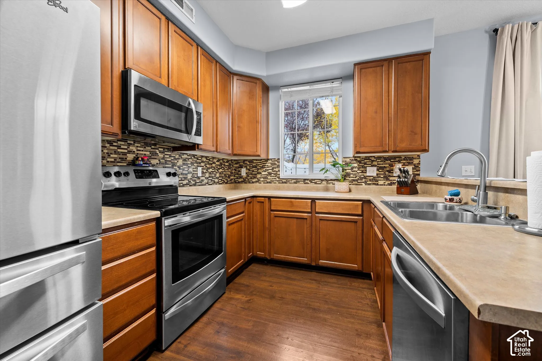 Kitchen featuring wood cabinetry, stainless steel appliances, decorative stone-patterned backsplash, solid-surface countertops, and wood-finish flooring