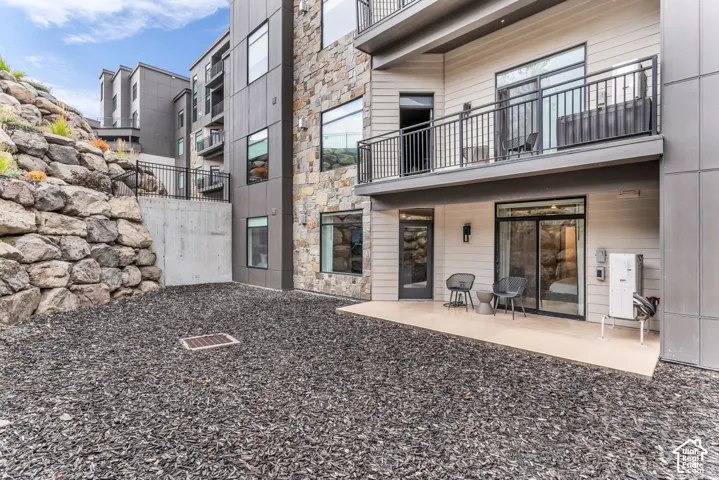Multi-story exterior featuring stone veneer, metal panel siding, a private balcony with metal railing, a ground-level concrete patio, and a tiered rock wall landscape