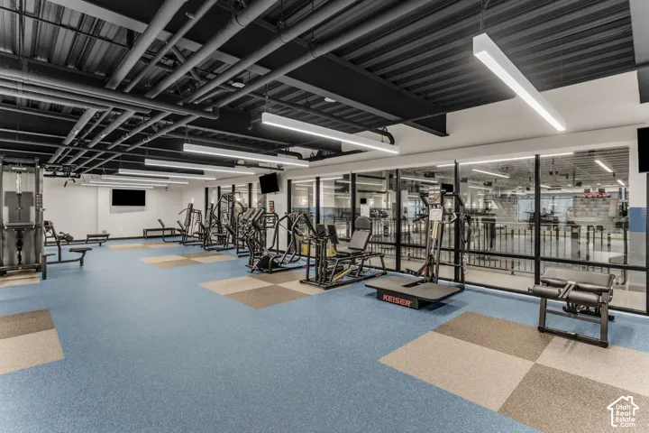 Industrial-style fitness center featuring an open-concept layout, exposed ceiling joists and ductwork, linear LED lighting, and rubberized tile flooring