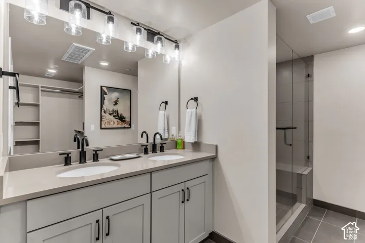 Dual-sink vanity with neutral countertop, matte black fixtures, full-width mirror with overhead glass lighting, and walk-in tiled shower with glass enclosure