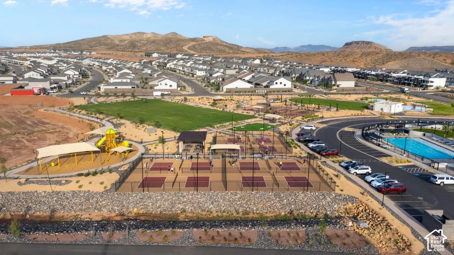 Aerial perspective of community recreational facilities featuring multiple pickleball courts, a swimming pool, a playground with shade structures, and an expansive grass lawn set against a mountain backdrop