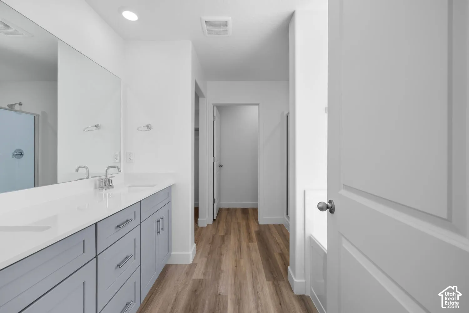 Bathroom featuring a double-sink vanity with light-gray cabinetry, solid-surface countertops, wood-finish flooring, and a large-format vanity mirror