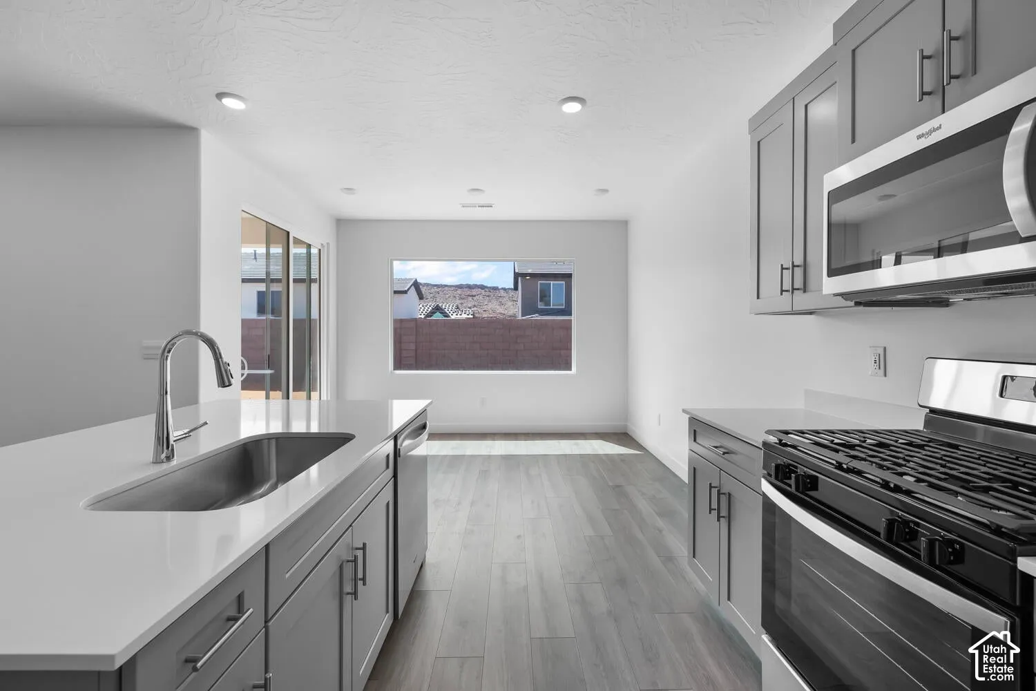 Kitchen featuring gray shaker-style cabinetry, quartz countertops, stainless steel undermount sink with high-arc faucet, stainless steel gas range, and wood-finish flooring