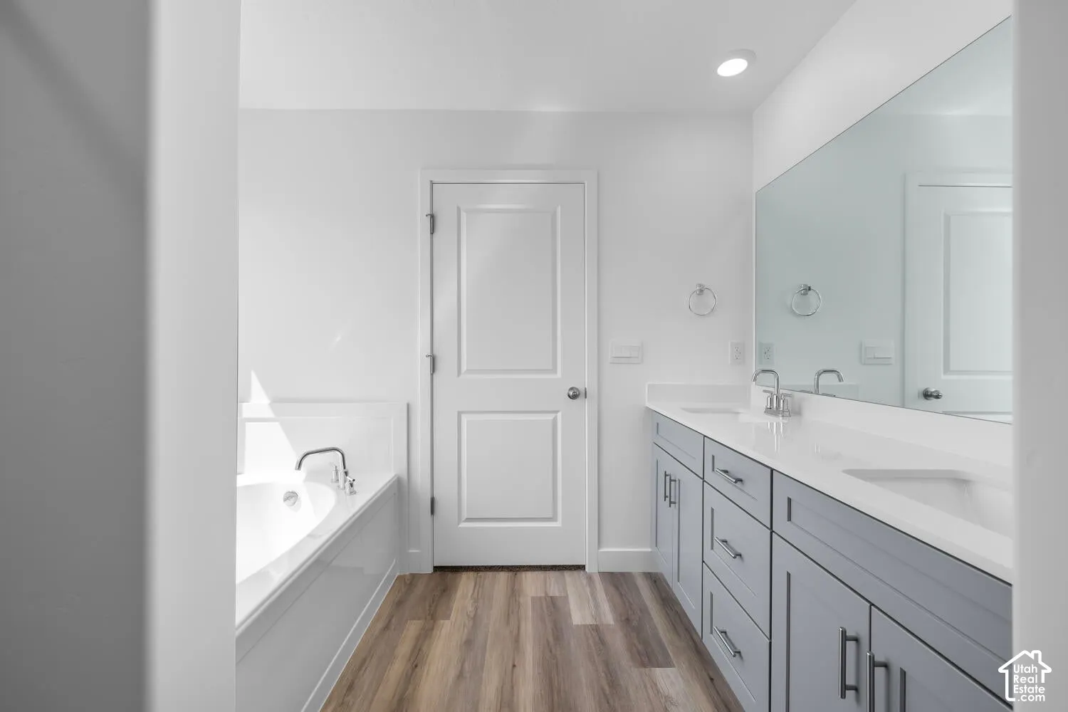 Dual-sink vanity with grey shaker-style cabinetry, white solid-surface countertop, recessed soaking tub, and wood-finish flooring