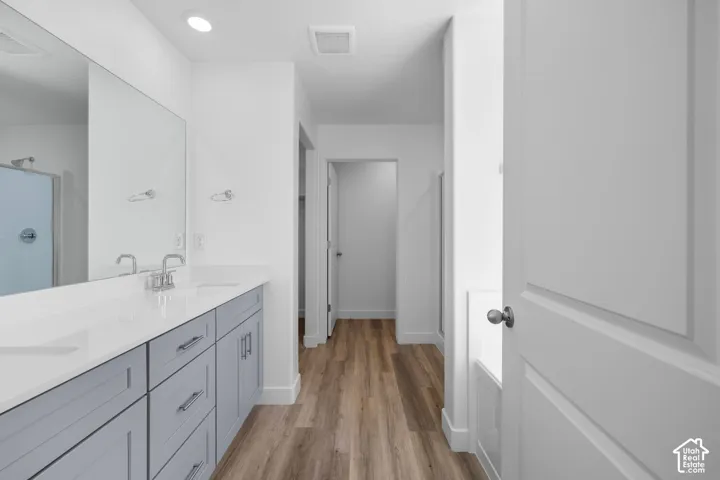 Bathroom featuring a double-sink vanity with light-gray cabinetry, solid-surface countertops, wood-finish flooring, and a large-format vanity mirror