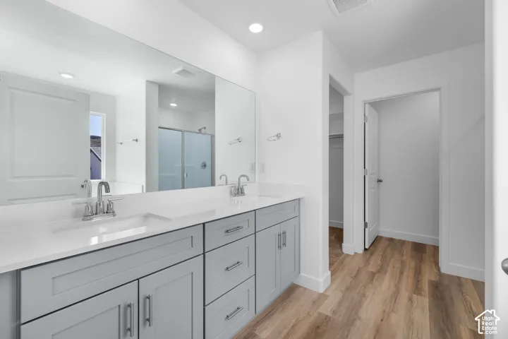 Dual-sink vanity with grey cabinetry, white solid-surface countertops, polished chrome fixtures, expansive wall-to-wall mirror, and wood-finish flooring