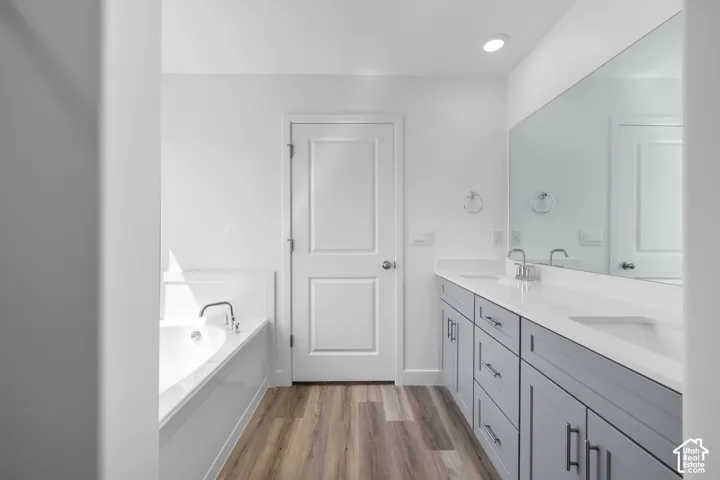 Dual-sink vanity with grey shaker-style cabinetry, white solid-surface countertop, recessed soaking tub, and wood-finish flooring