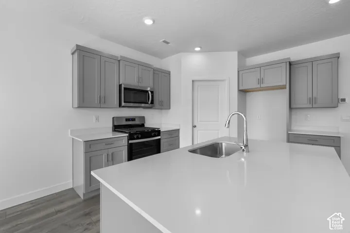 Kitchen featuring gray shaker-style cabinetry, white solid-surface countertops, a center island with a stainless steel undermount sink, a gas range, and wood-finish flooring