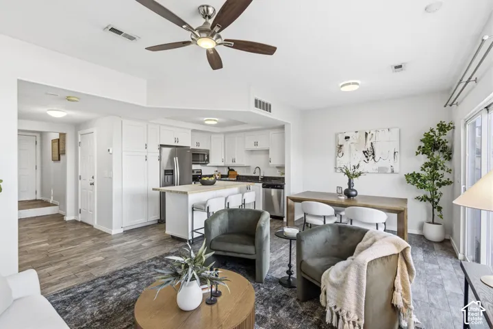 Open-concept living space featuring wood-finish flooring, white shaker-style cabinetry, stainless steel kitchen appliances, a kitchen island, and a ceiling fan with integrated lighting