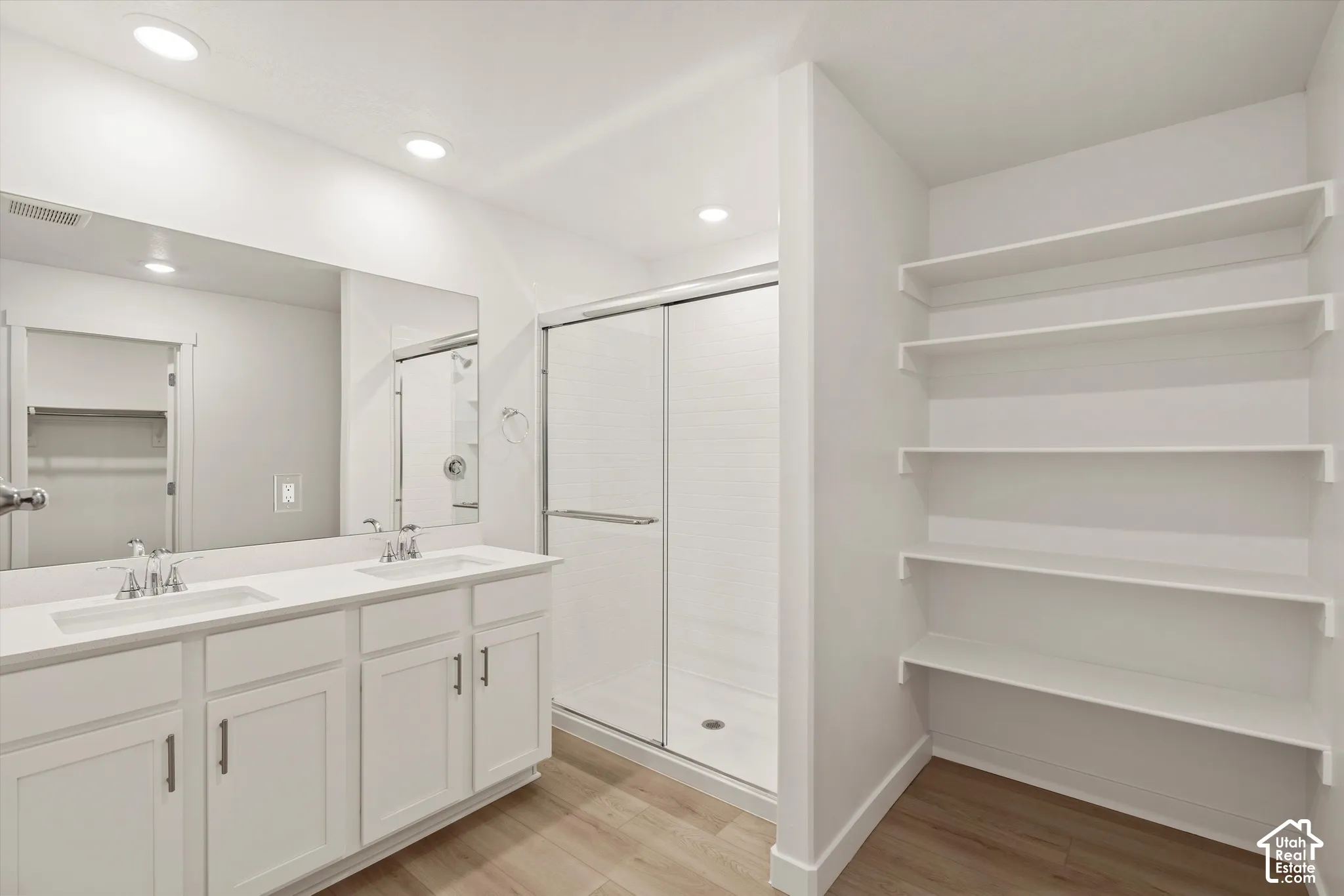 **Disclaimer** Interior package & colors may vary.  Dual-sink vanity with white cabinetry and countertop, frameless glass shower enclosure, recessed lighting, and built-in shelving with wood-finish flooring