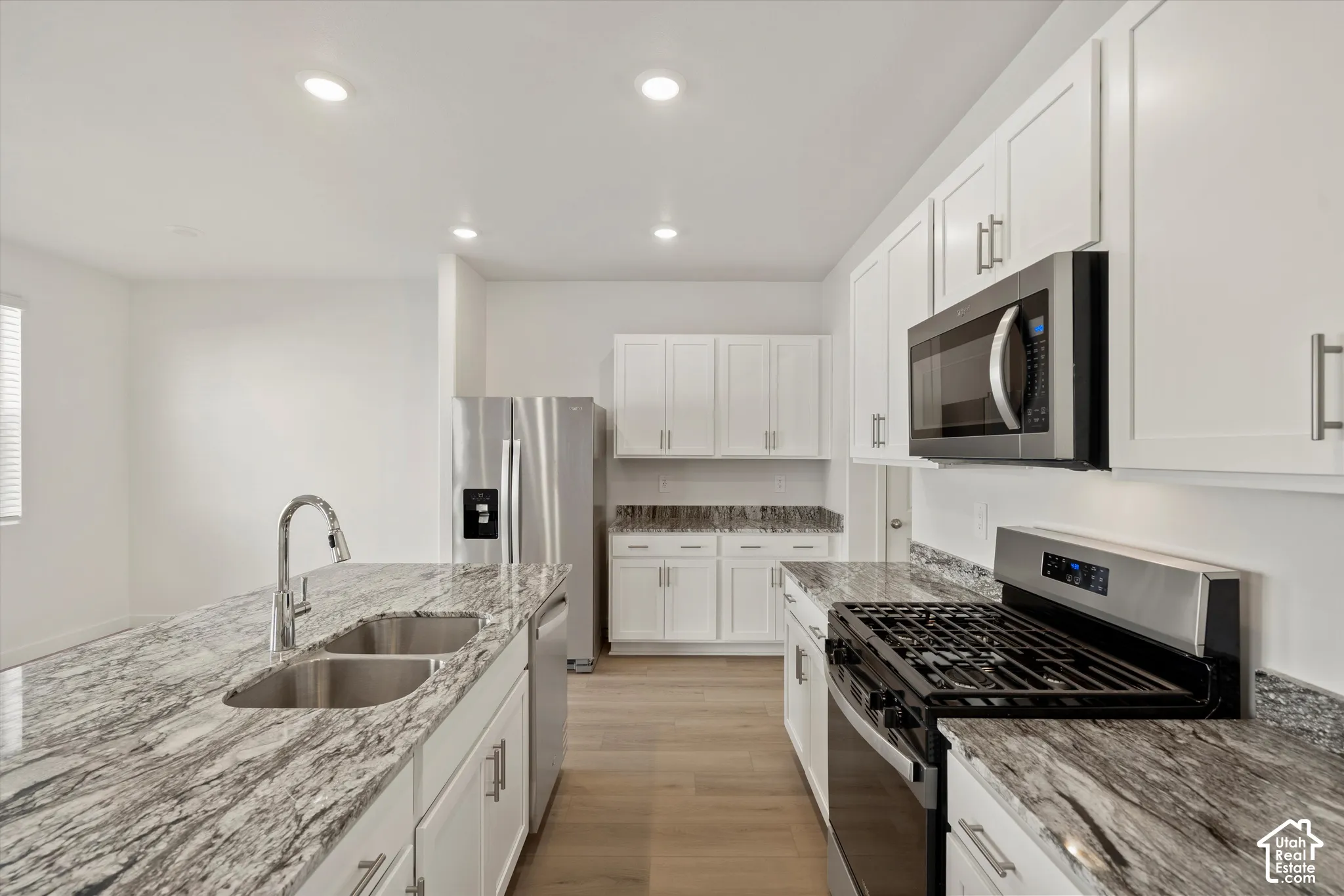 **Disclaimer** Interior package & colors may vary.  Kitchen featuring white shaker-style cabinetry, stone countertops, stainless steel appliances, a central island with an undermount double-basin sink, and wood-finish flooring