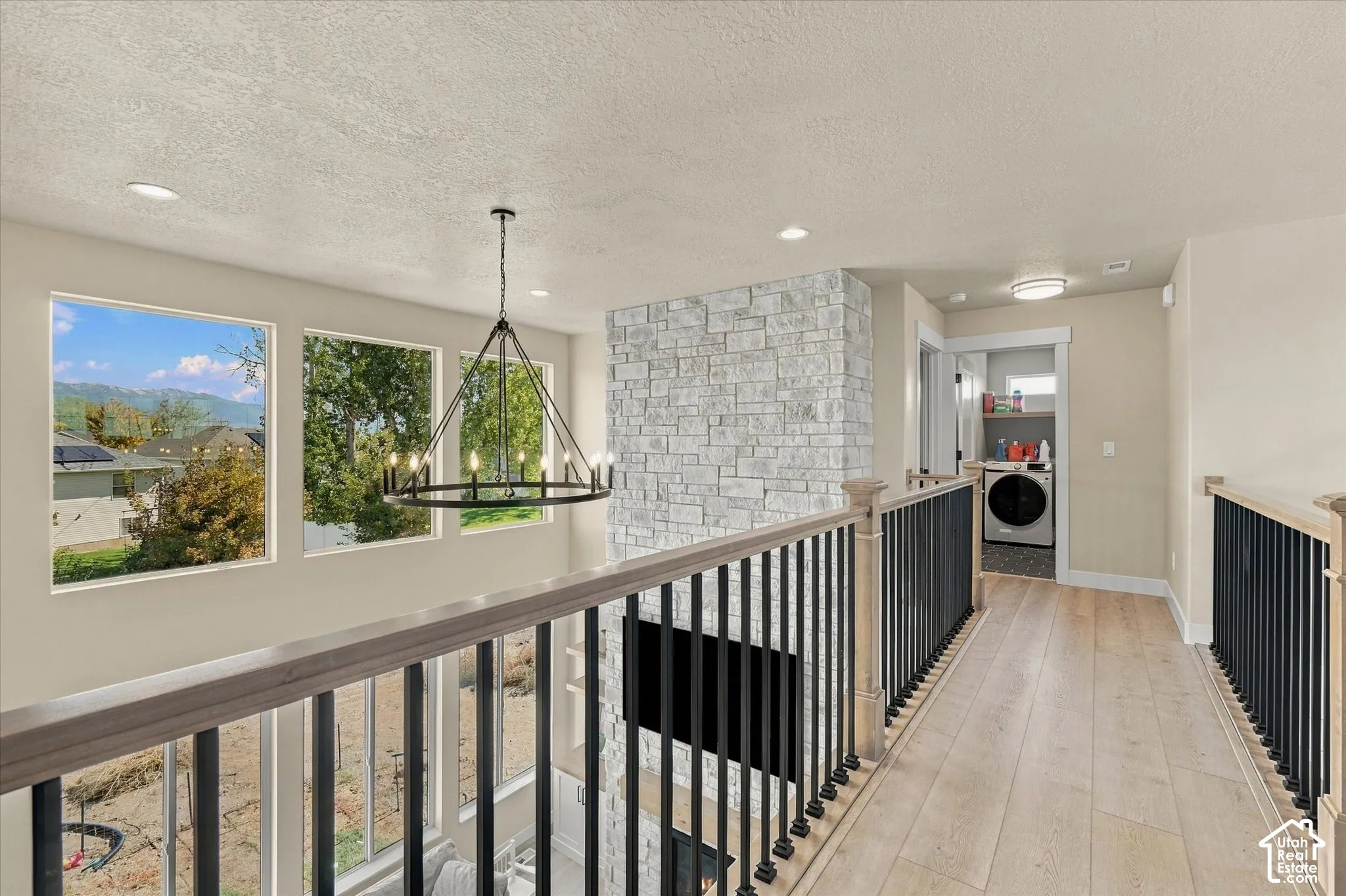 Elevated landing featuring wood-finish flooring, metal spindle railing, floor-to-ceiling stone-faced chimney, modern multi-arm chandelier, and recessed lighting
