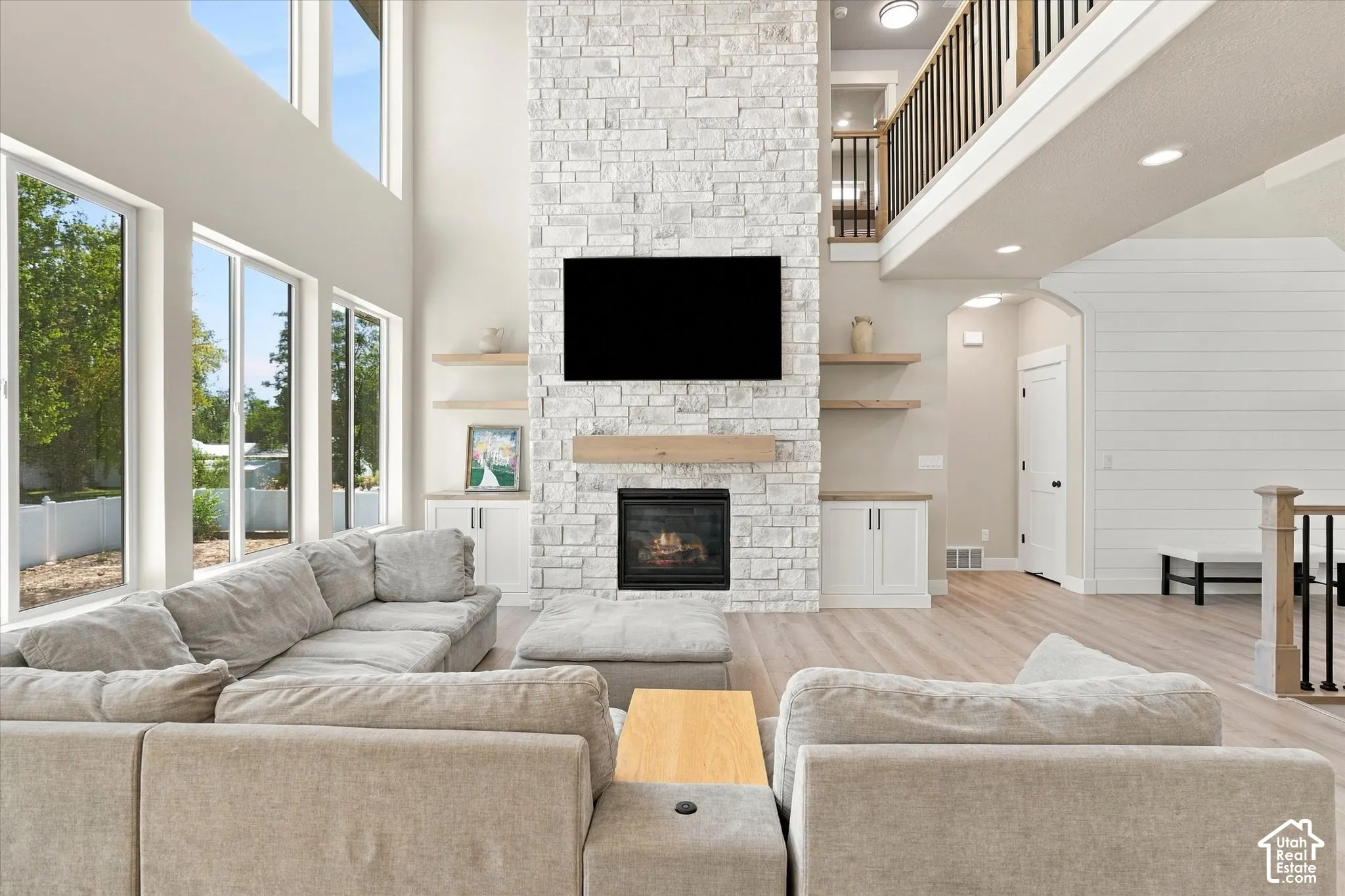 Double-height living area featuring floor-to-ceiling windows, stacked stone fireplace, built-in cabinetry with floating shelving, and wood-finish flooring