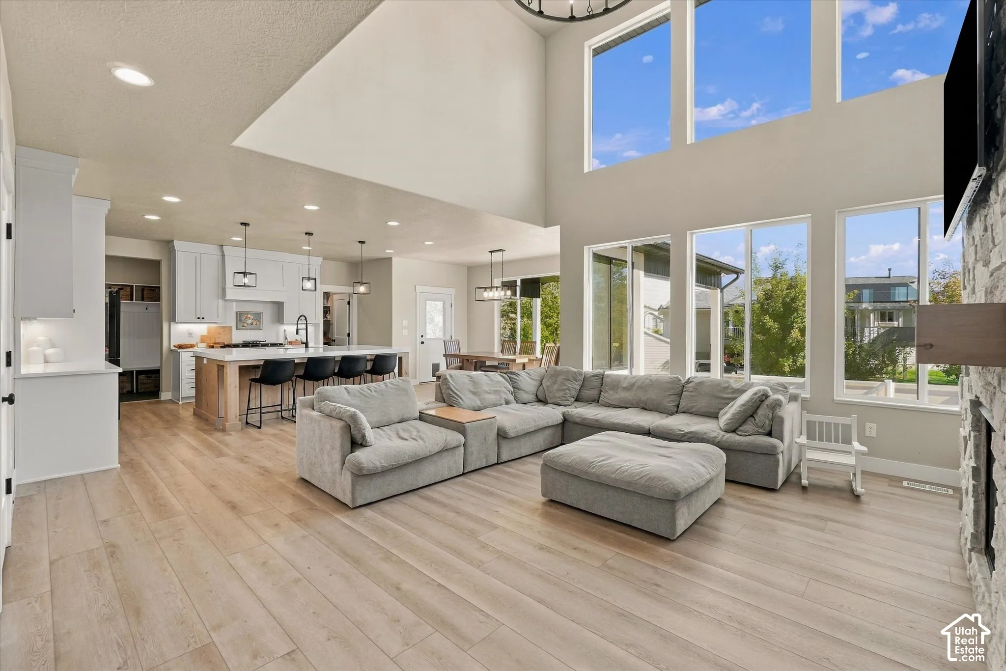 Open-concept living space featuring vaulted ceilings, floor-to-ceiling windows, wood-finish flooring, a stone-surround fireplace, and a kitchen with an oversized island