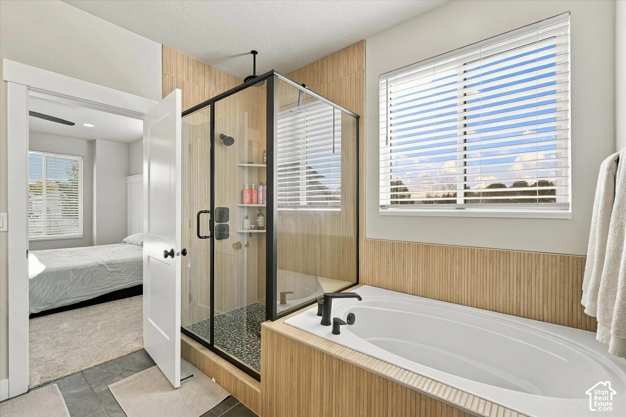 Glass-enclosed shower with a matte black frame, deep soaking tub, vertical slat-style wall tile, matte black fixtures, and window with slatted blinds