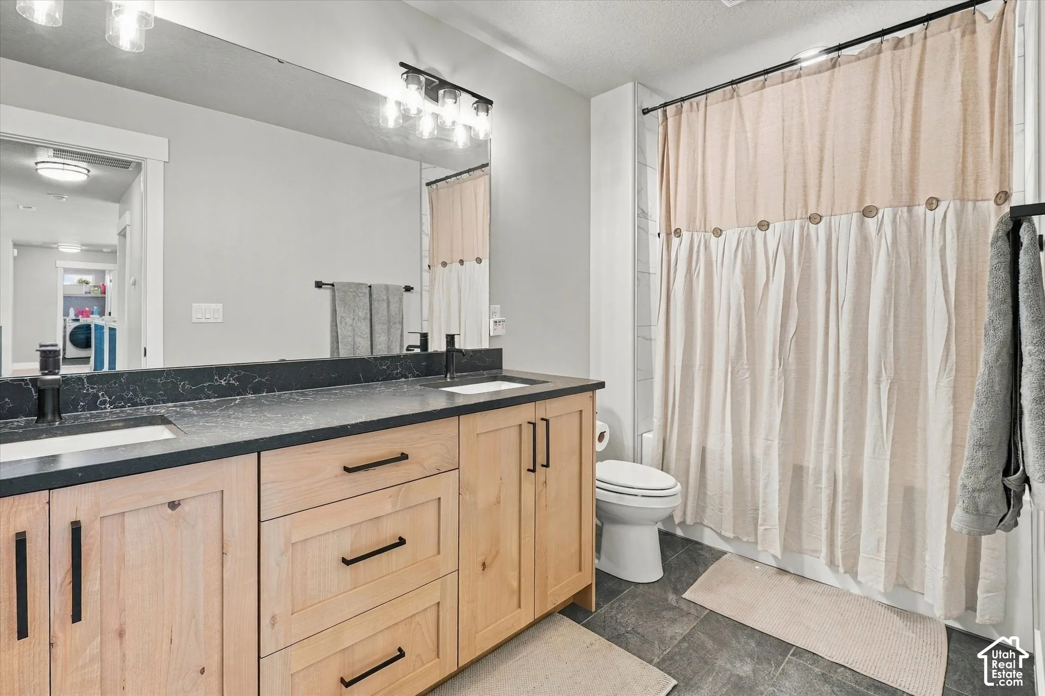 Double vanity featuring light wood-finish cabinetry, dark contrasting countertops with integrated sinks, matte black hardware, and an expansive wall-mounted mirror