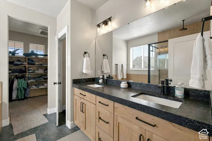 Dual-sink vanity with matte black hardware, stone-finish countertops, wall-to-wall mirror, and light wood cabinetry, featuring tile flooring and walk-in storage with built-in shelving