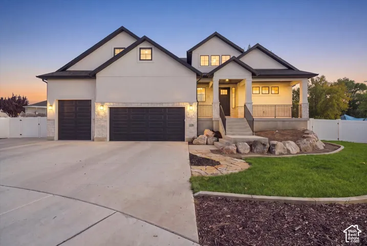 Two-story exterior featuring a light-toned stucco finish, multi-gabled roofline, stone veneer accents, three-car garage, and a covered front porch with iron railings