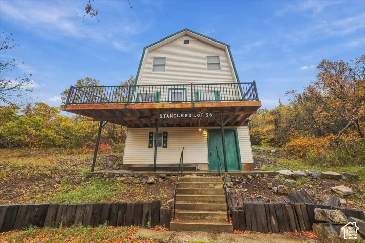 Two-story gambrel-roof structure featuring horizontal siding, a cantilevered upper-level deck with metal railing, concrete entry stairs with metal handrails, and a retaining wall constructed of stacked timber sleepers