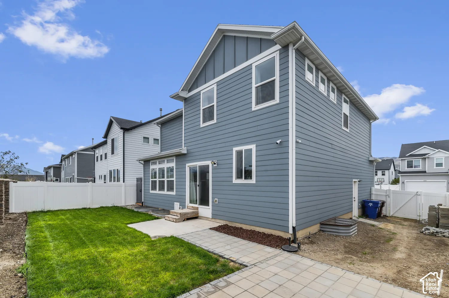 Exterior featuring blue horizontal siding, vertical board-and-batten gable accents, a white vinyl privacy fence, a concrete patio, and paver stone walkway